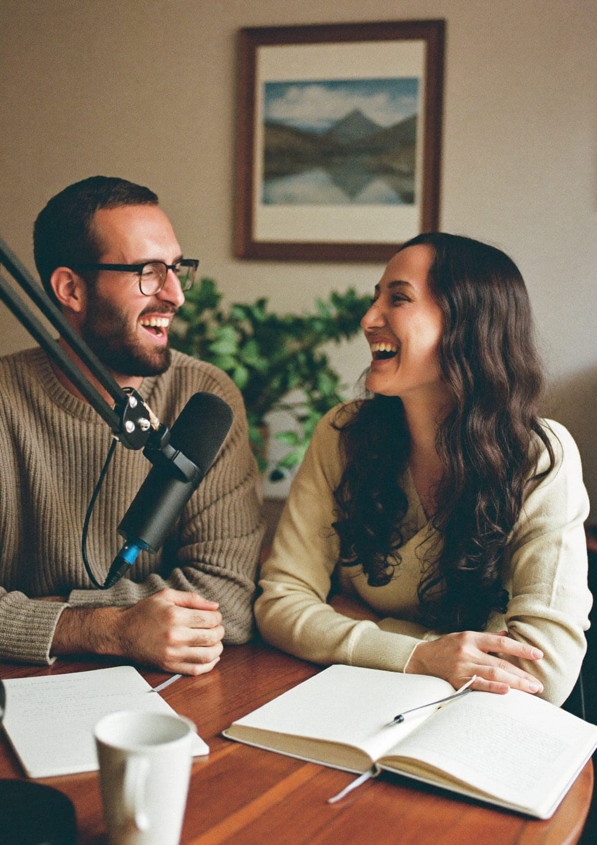 Two people laughing mid-conversation at a table with a podcast microphone